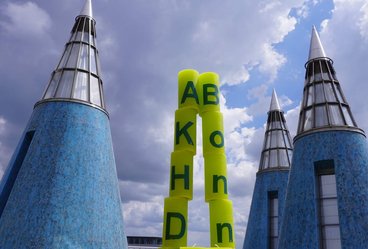 Das Foto zeigt die Jubiläumskerzen als spitzen Kegel aufgestellt. Links stehen die Kerzen mit den Buchstaben A, K, H, D übereinander, rechts die mit dem Schriftzug "Bonn". Unten stehen die Kerzen mit der "10" davor. Im Hintergrund sind die blauen Kegel auf dem Dach der Bundeskunsthalle zu sehen.