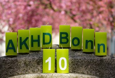Das Foto zeigt unsere zehn grünen Jubiläumskerzen mit der Aufschrift "AKHD Bonn 10" auf dem Kopf eines Steinsockels in der Bonner Heerstraße unter dem rosafarbenen Dach der Kirschblüten.
