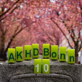 Das Foto zeigt unsere zehn grünen Jubiläumskerzen mit der Aufschrift "AKHD Bonn 10" auf dem Kopf eines Steinsockels in der Bonner Heerstraße. Darüber wölbt sich das rosafarbene Dach der Kirschblüten.